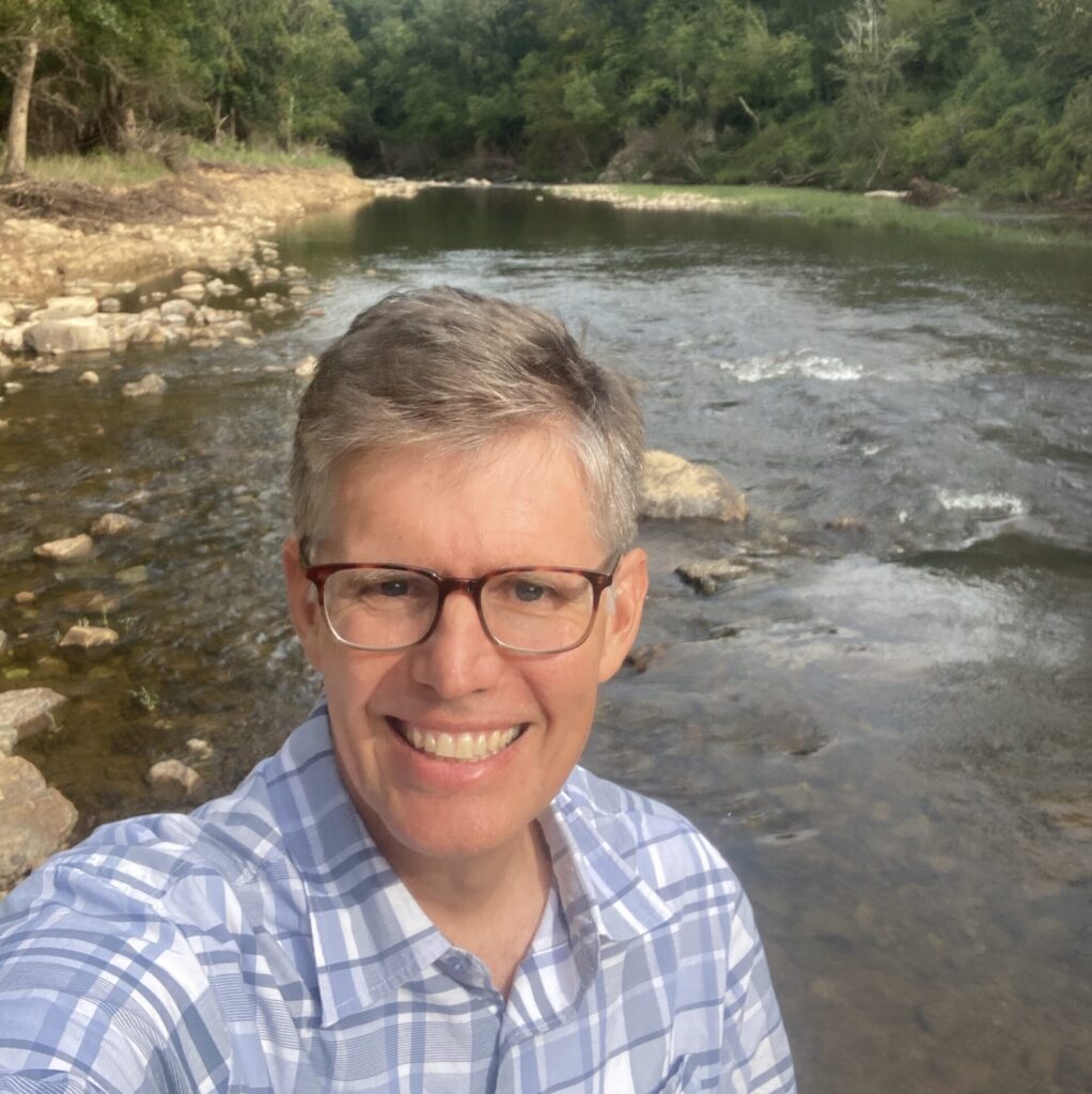 Chest-up photo of Chris Dreps wearing a blue plaid shirt in front of a stretch of the Eno.
