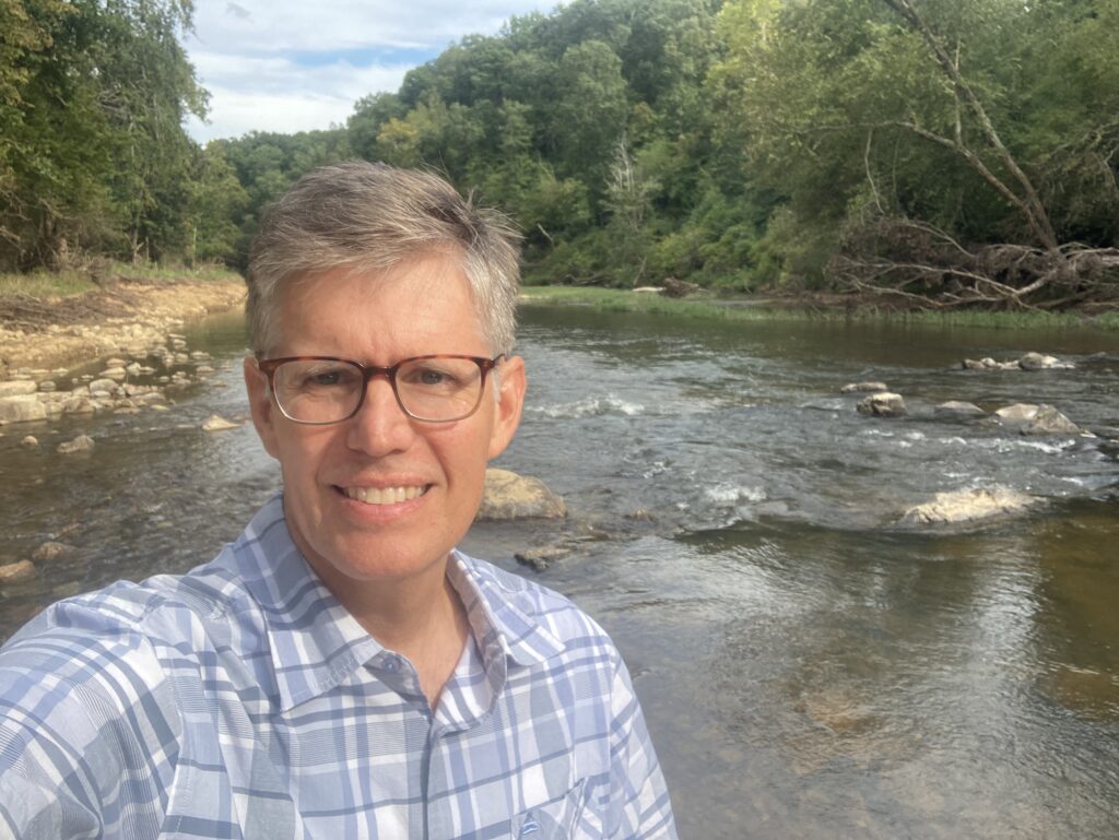 Chest-up photo of Chris Dreps wearing a blue plaid shirt in front of a stretch of the Eno.