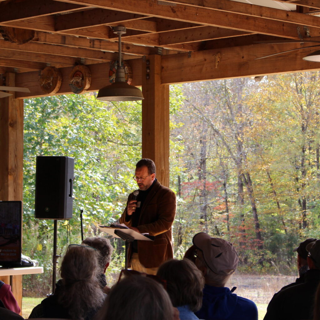 Photo of Ryan Fehrman speaking in front of members at the 2025 Annual Meeting, with the autumnal Confluence Natural Area in the background.