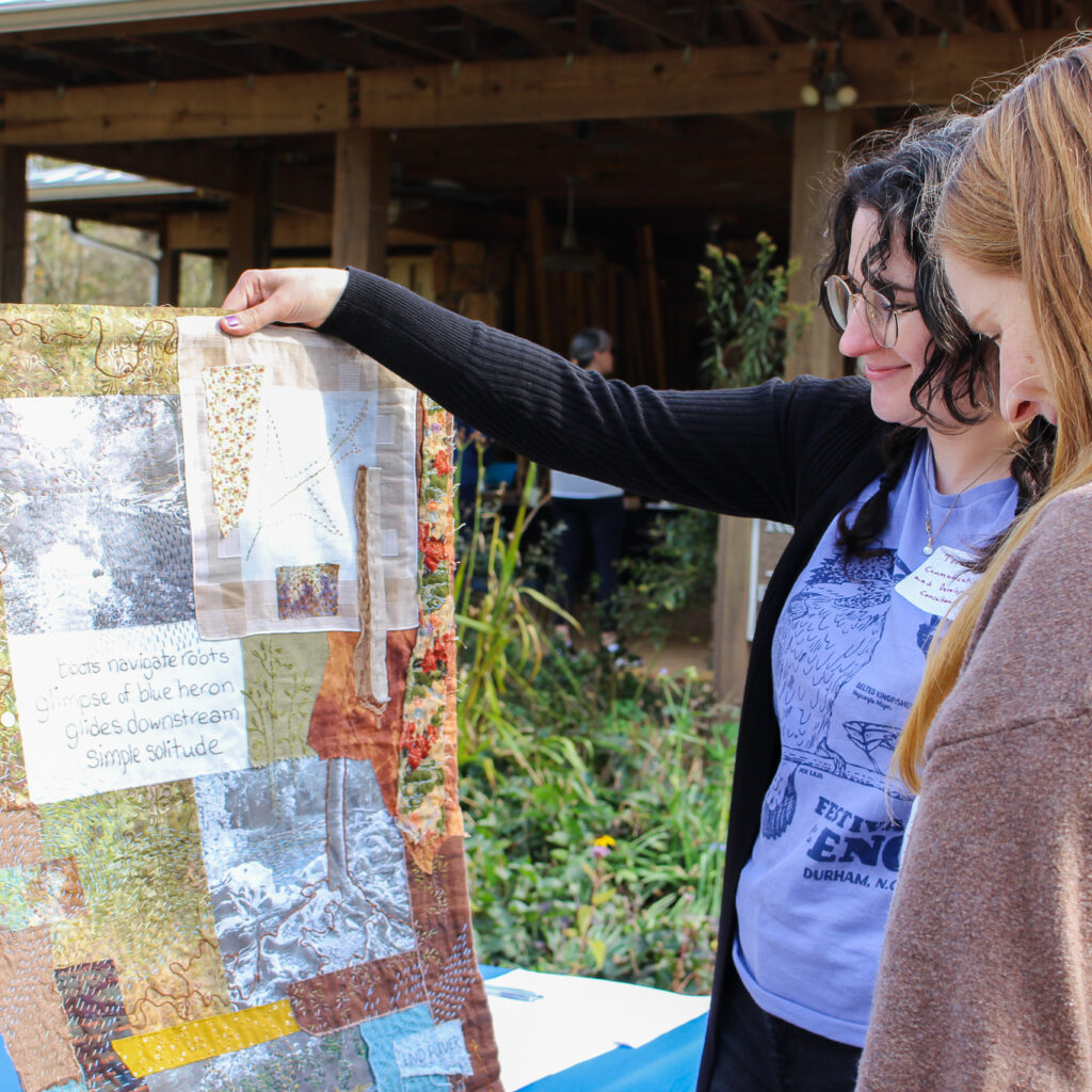 ERA staff looking at an art quilt by Carol Gunther-Mohr. The quilt is made up of colorful fabric stitched together with photos of the Eno, with a haiku by Carol. The haiku reads: "boots navigate roots, glimpse of blue heron glides downstream, simple solitude"