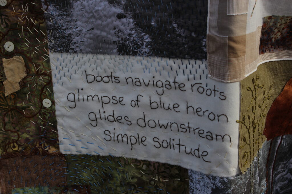 Close-up of haiku on art quilt by Carol Gunther-Mohr. Reads "boots navigate roots, glimpse of blue heron glides downstream, simple solitude."